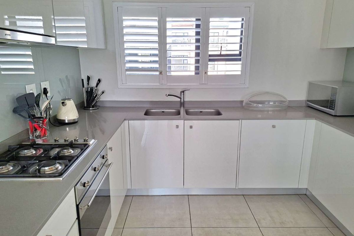 Modern kitchen featuring white gloss cabinetry, stone countertops, a gas hob, built-in oven, and plantation shutters over the window.