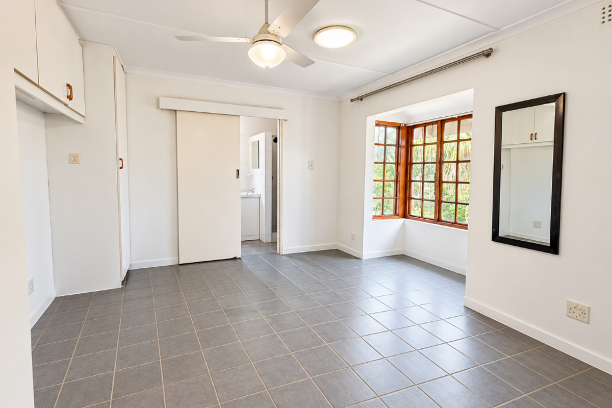 A bright, empty bedroom featuring tiled flooring, built-in cupboards, a ceiling fan, and a bay window with views of the garden.