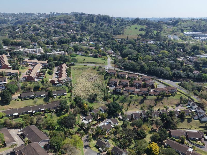 An aerial view of a residential area showing a vacant plot of land surrounded by established housing developments and lush greenery.