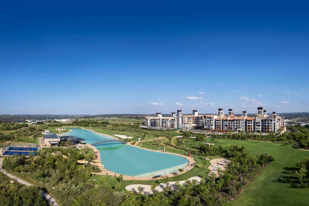 Aerial view of a modern apartment complex featuring a large man-made lagoon, pedestrian bridge, landscaped grounds, and recreational sports facilities.