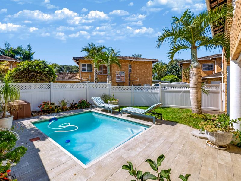 A well-maintained private backyard featuring a swimming pool, tiled patio area, and perimeter vinyl fencing, surrounded by established palm trees.