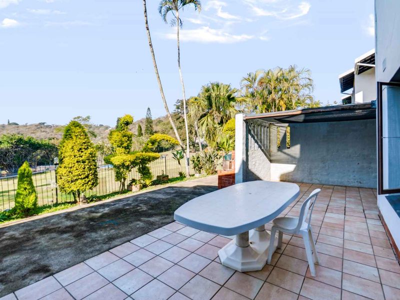 Spacious covered patio featuring tiled flooring and a built-in brick braai, overlooking a landscaped garden area with scenic views.