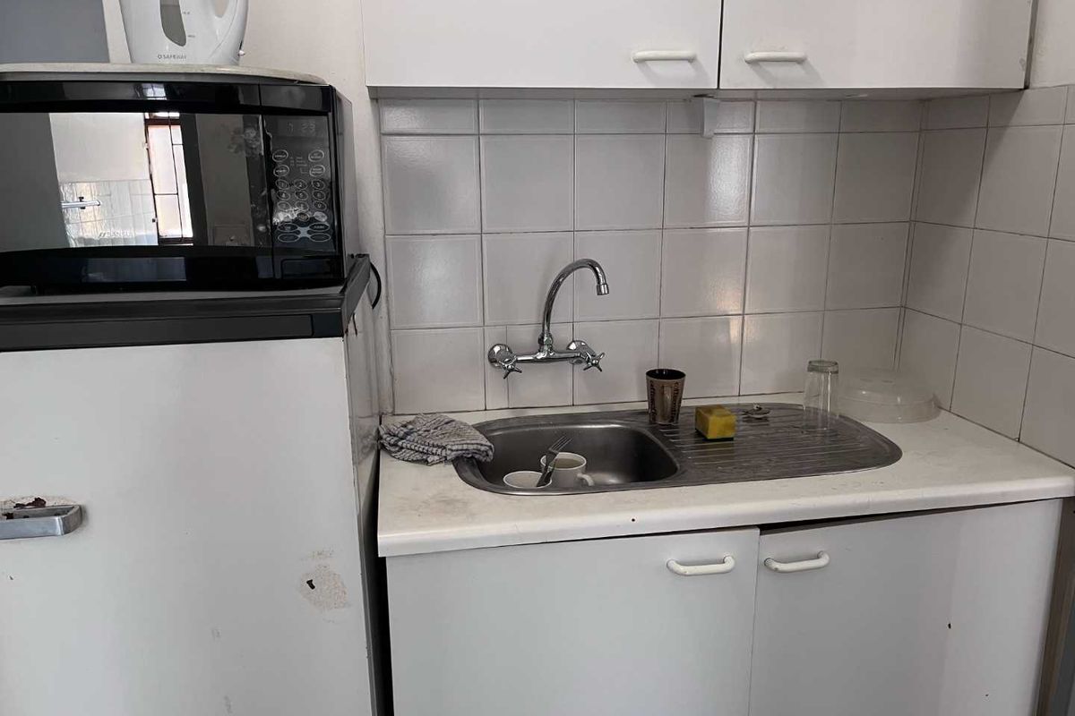 A compact kitchenette area featuring a stainless steel sink, wall-mounted faucet, tiled backsplash, and white cabinetry.