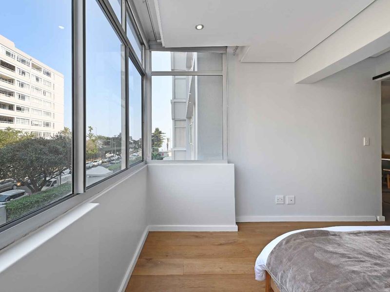 A bright bedroom featuring expansive floor-to-ceiling windows with urban views, modern recessed lighting, and high-quality timber flooring.