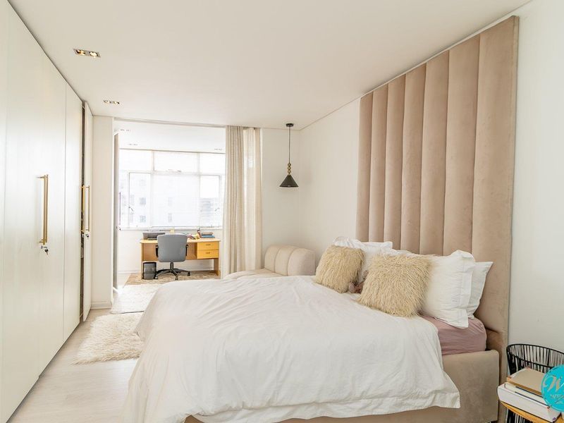 A modern, well-maintained bedroom featuring extensive built-in cabinetry, a floor-to-ceiling upholstered headboard panel, and recessed lighting, with an adjoining study nook.