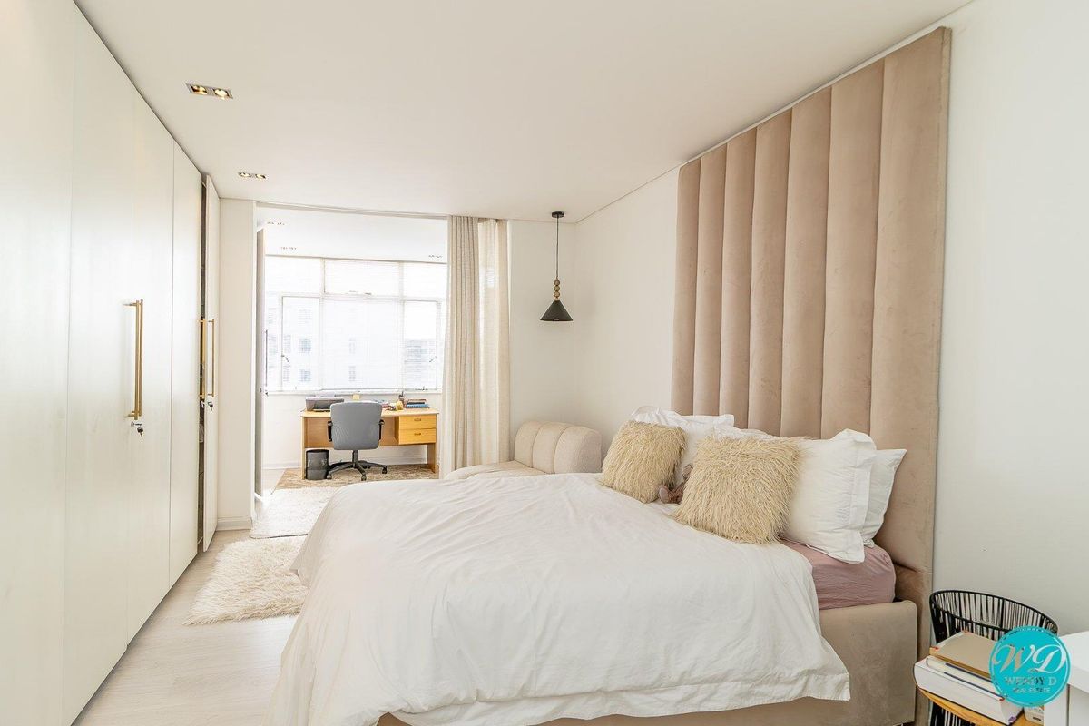 A modern, well-maintained bedroom featuring extensive built-in cabinetry, a floor-to-ceiling upholstered headboard panel, and recessed lighting, with an adjoining study nook.