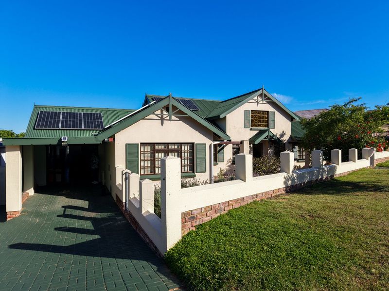 Front elevation of a single-story house featuring a green metal roof, solar panels, a paved driveway, and a perimeter wall with gated entrance.