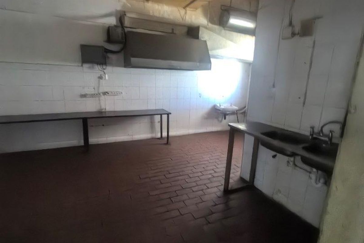A utilitarian commercial-style kitchen space featuring stainless steel prep surfaces, a sink, an extractor hood, tiled walls, and durable brick flooring.
