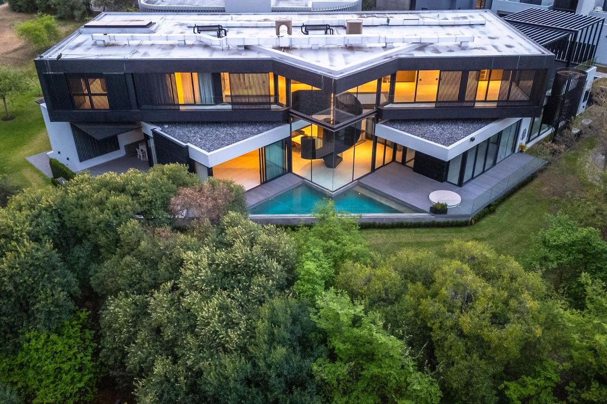 Aerial view of a modern luxury residence featuring a unique architectural design, glass facade, private swimming pool, and expansive decking surrounded by lush greenery.