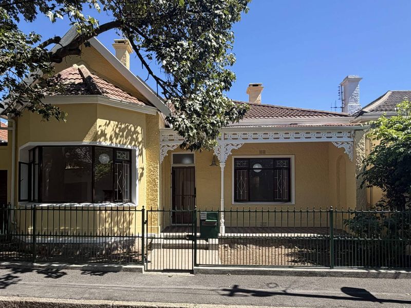 Charming yellow stucco house featuring a front veranda with decorative fretwork, a bay window, and a secure metal fence.