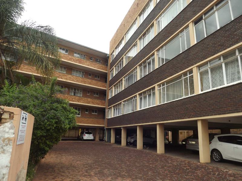 Exterior view of a multi-story brick apartment building featuring a paved driveway and covered ground-level parking area.