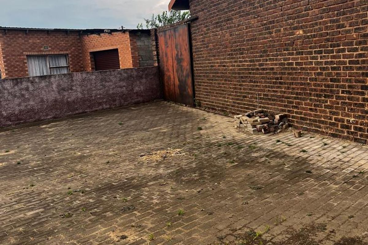 A rear exterior courtyard featuring brick paving, exposed brick walls, and a metal gate, requiring maintenance and landscaping to improve its overall appearance.