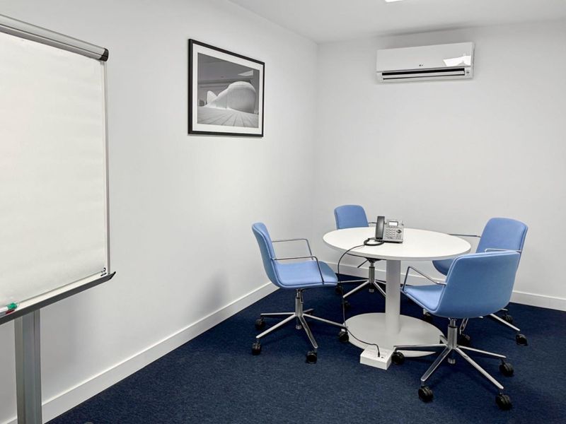 A clean and functional office meeting room featuring blue carpeting, a wall-mounted air conditioning unit, and neutral white walls.