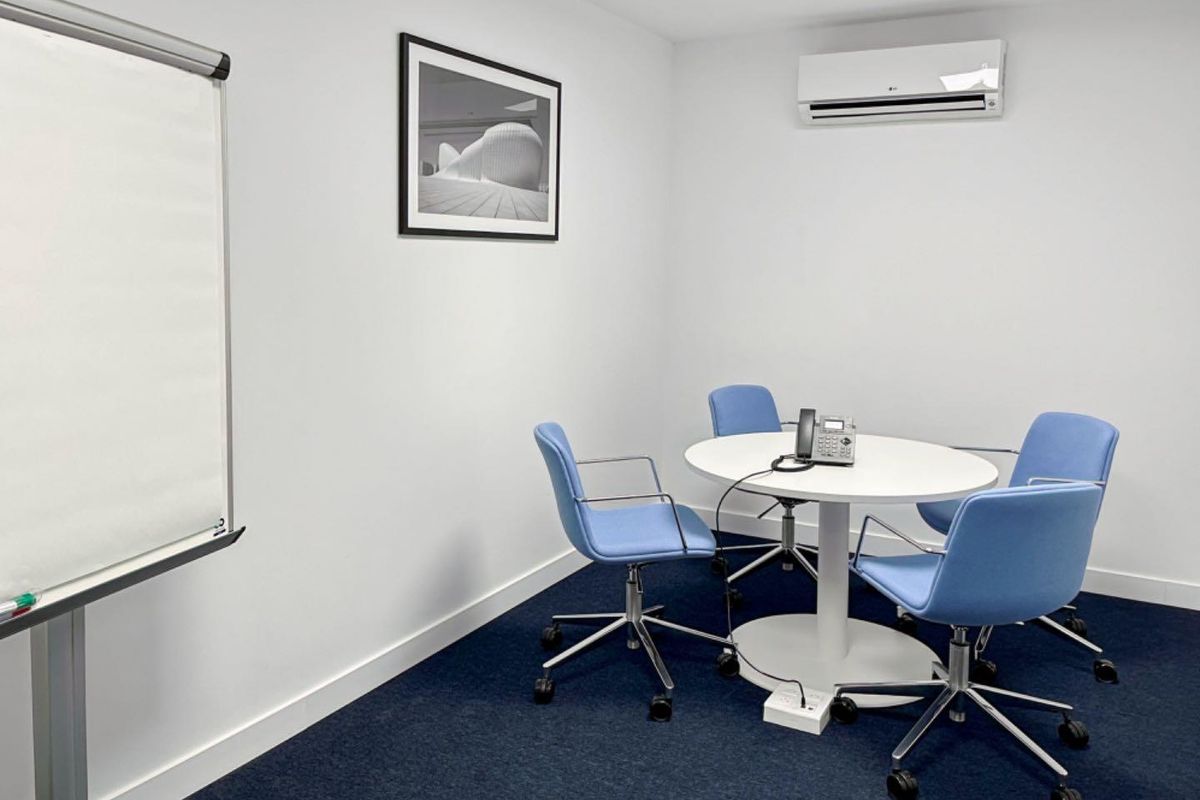 A clean and functional office meeting room featuring blue carpeting, a wall-mounted air conditioning unit, and neutral white walls.