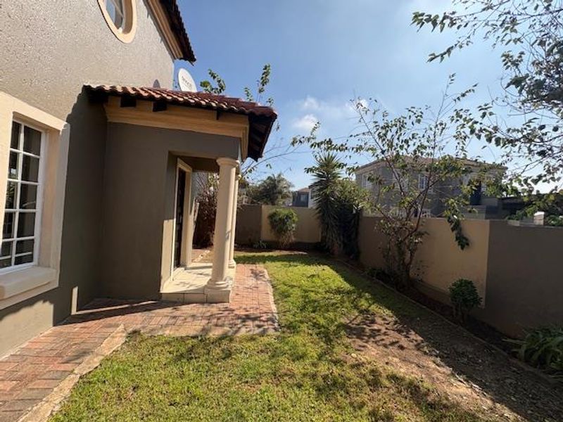 A rear exterior view of the townhouse featuring a paved patio area, covered entrance with columns, and a private garden enclosed by boundary walls.