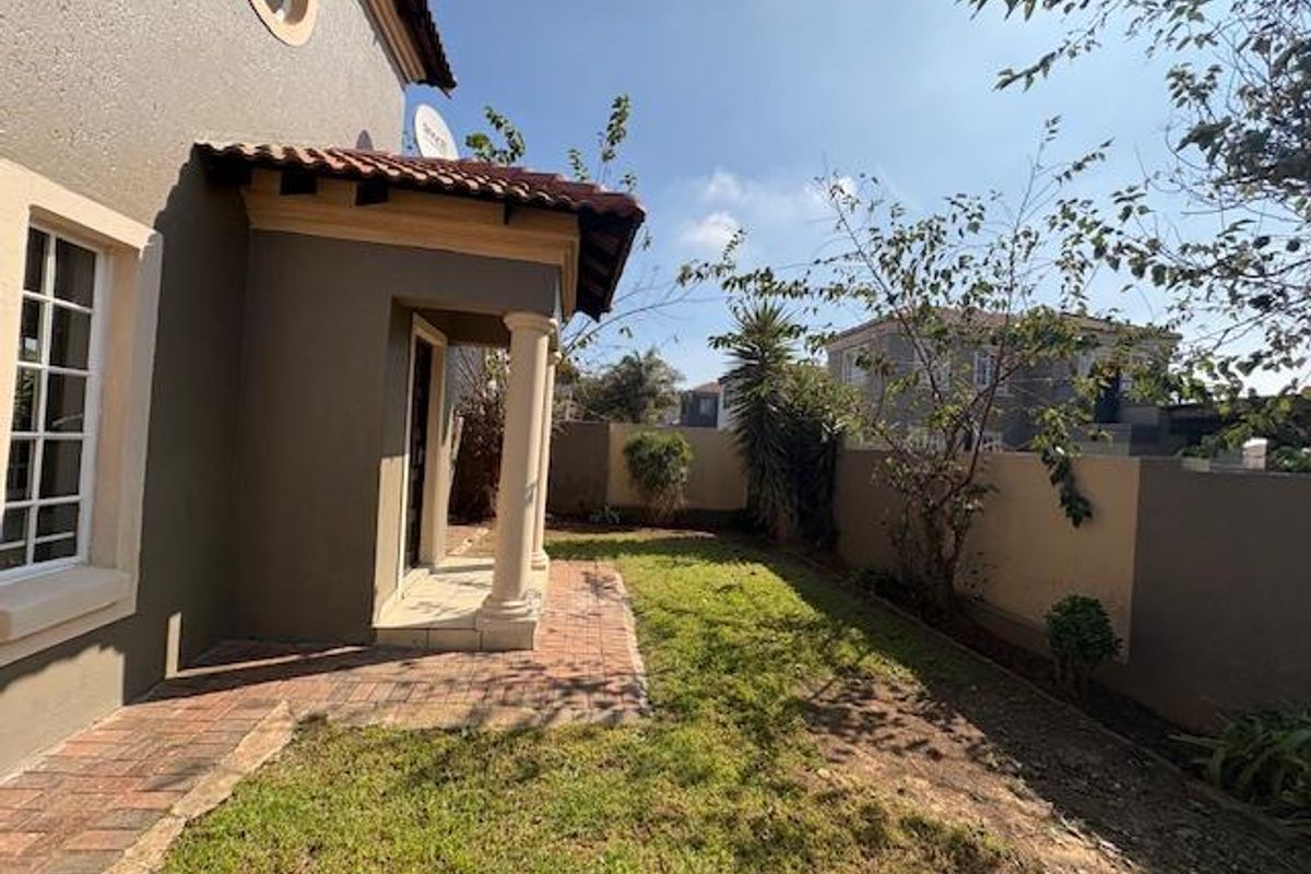 A rear exterior view of the townhouse featuring a paved patio area, covered entrance with columns, and a private garden enclosed by boundary walls.