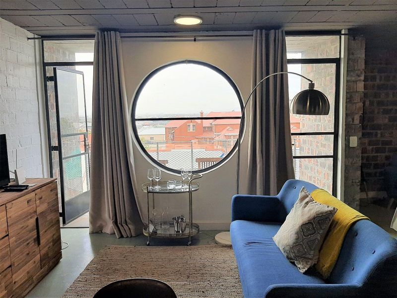 A stylish living room featuring exposed brick walls, an industrial concrete ceiling, and a unique circular window with access to a private balcony.