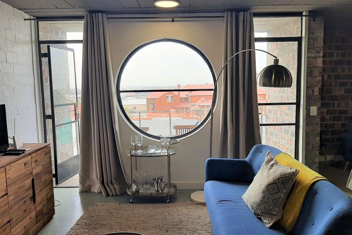 A stylish living room featuring exposed brick walls, an industrial concrete ceiling, and a unique circular window with access to a private balcony.