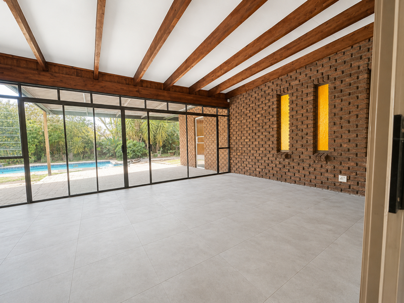 Spacious enclosed patio featuring exposed timber beams, a rustic brick feature wall with amber glass accents, and large sliding doors overlooking the pool area.
