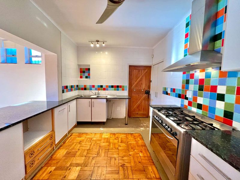 A functional kitchen featuring parquet flooring, a gas stove with stainless steel extractor, and a colorful tiled splashback with ample built-in cabinetry.