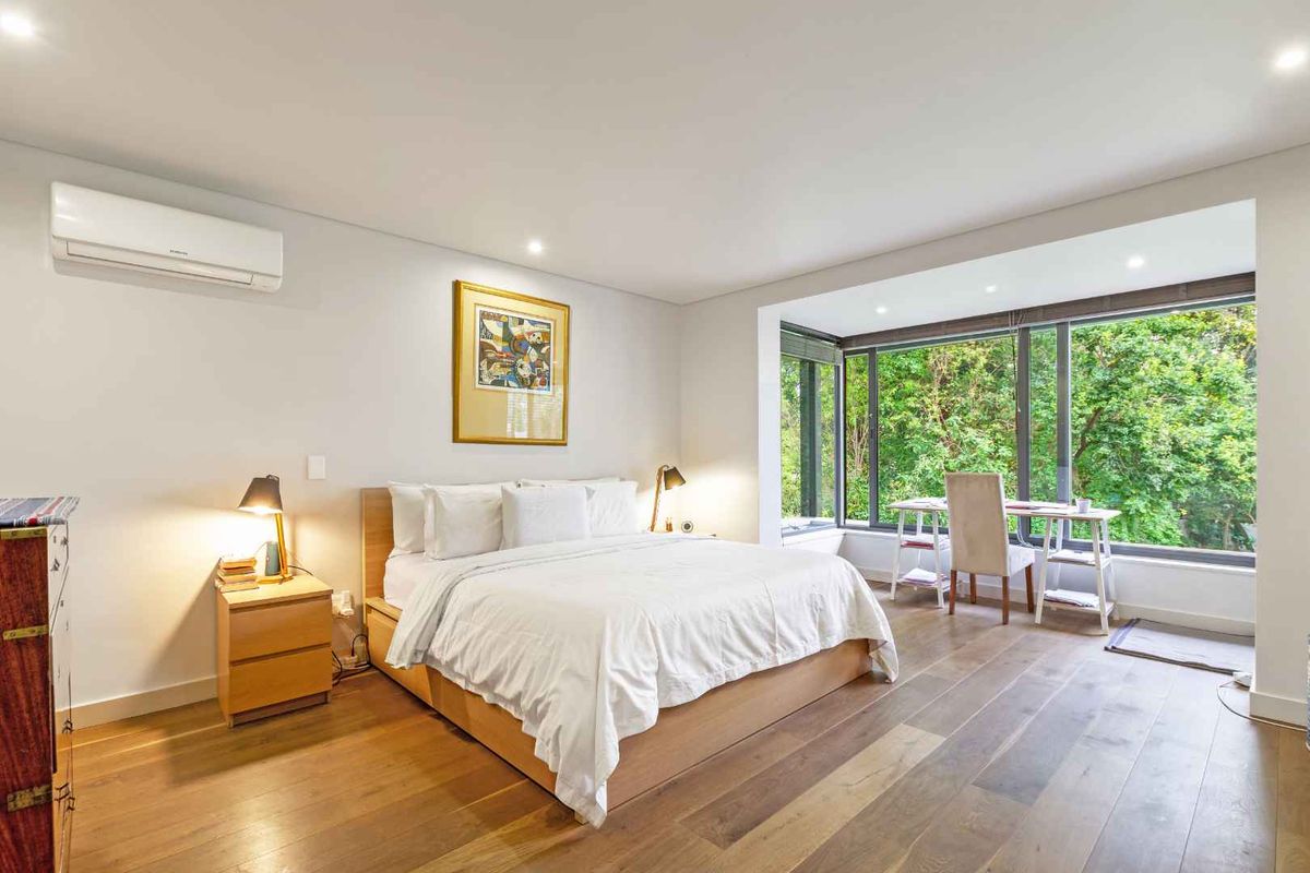 Spacious bedroom featuring hardwood flooring, recessed lighting, a wall-mounted air conditioning unit, and large floor-to-ceiling windows overlooking lush greenery.