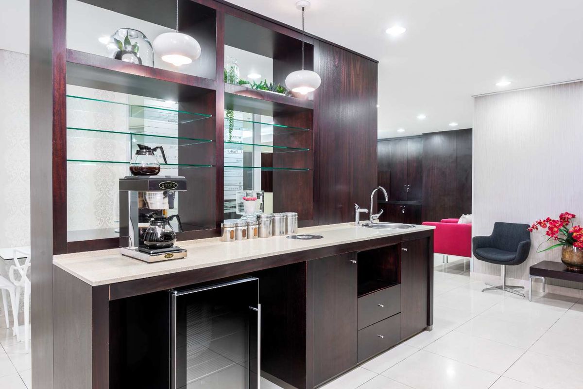 A modern office kitchenette featuring dark wood cabinetry, integrated shelving, a stone countertop, a stainless steel sink, and a built-in under-counter beverage fridge.