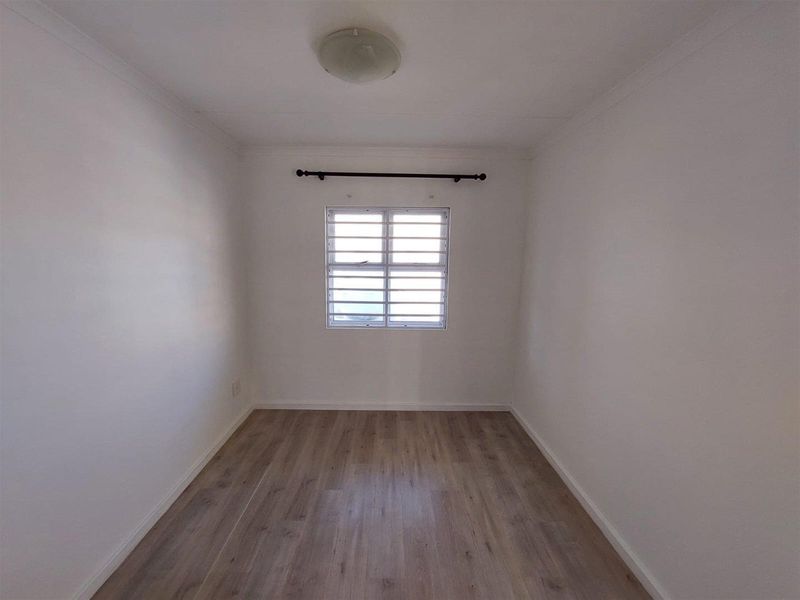 A clean, empty bedroom featuring light-toned laminate flooring, white walls, and a window fitted with security bars.