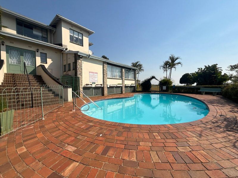 A well-maintained outdoor pool area featuring circular brick paving, stone-clad exterior walls, and a secure metal pool fence.