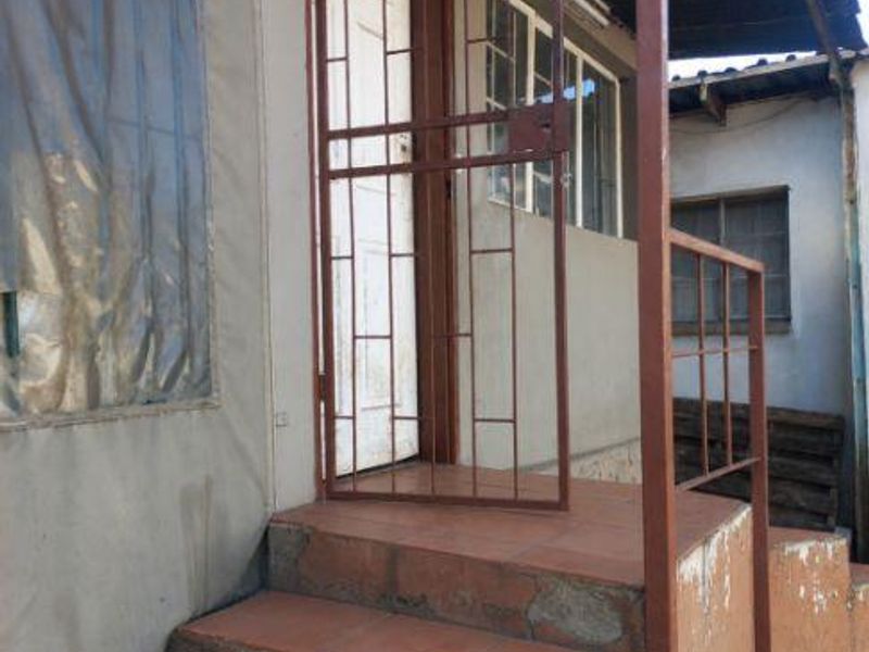 Exterior entrance area featuring tiled steps, a metal security gate, and a corrugated roof structure showing signs of wear and weathering.