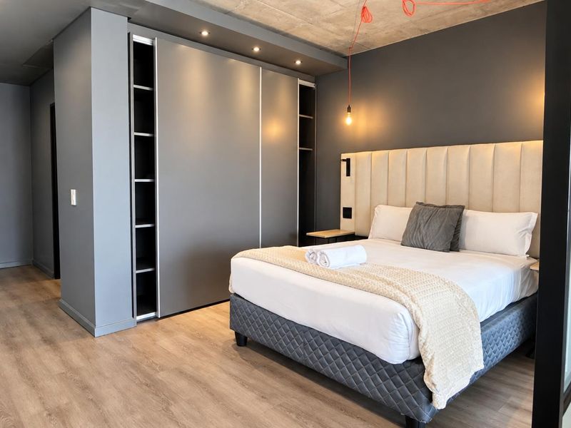 Modern bedroom featuring exposed concrete ceilings, laminate flooring, built-in wardrobes with integrated lighting, and a sleek glass partition.