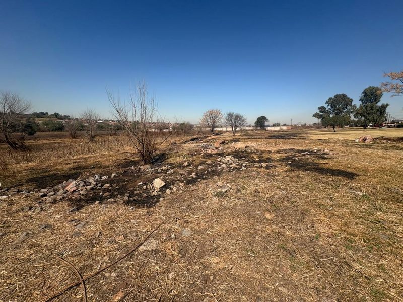 A view of a vacant residential plot featuring dry, scorched grass and uneven terrain under a clear blue sky.