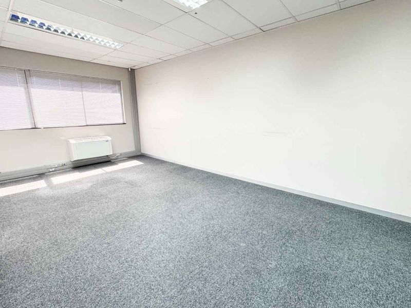A vacant, well-lit office space featuring neutral carpeting, a suspended ceiling with recessed lighting, and a wall-mounted HVAC unit.
