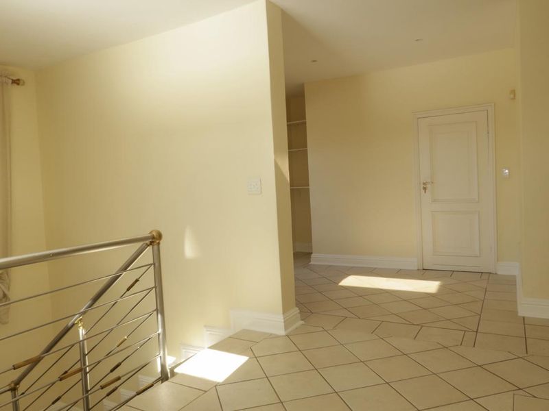 A bright, tiled hallway featuring a modern stainless steel balustrade and neutral wall finishes, leading to an adjacent room with built-in shelving.