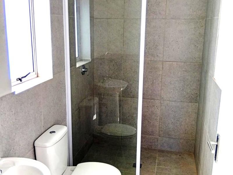 A clean, functional bathroom featuring a glass-enclosed shower, pedestal basin, and toilet with neutral grey wall and floor tiling.