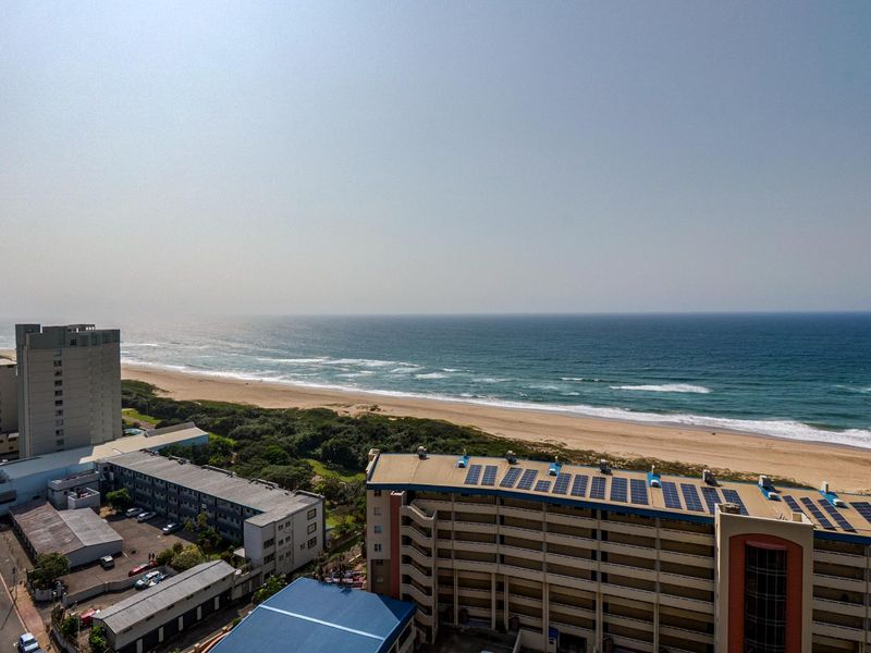 Elevated coastal view overlooking the ocean, sandy beach, and surrounding apartment complex with rooftop solar panel installations.