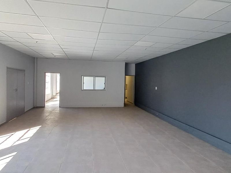 Spacious, open-plan industrial office area featuring a suspended ceiling, tiled flooring, large windows for natural light, and a modern dark accent wall.