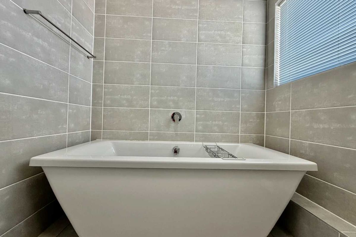 A modern bathroom featuring a clean, white freestanding bathtub set against neutral grey tiled walls with a wall-mounted mixer and a window blind.