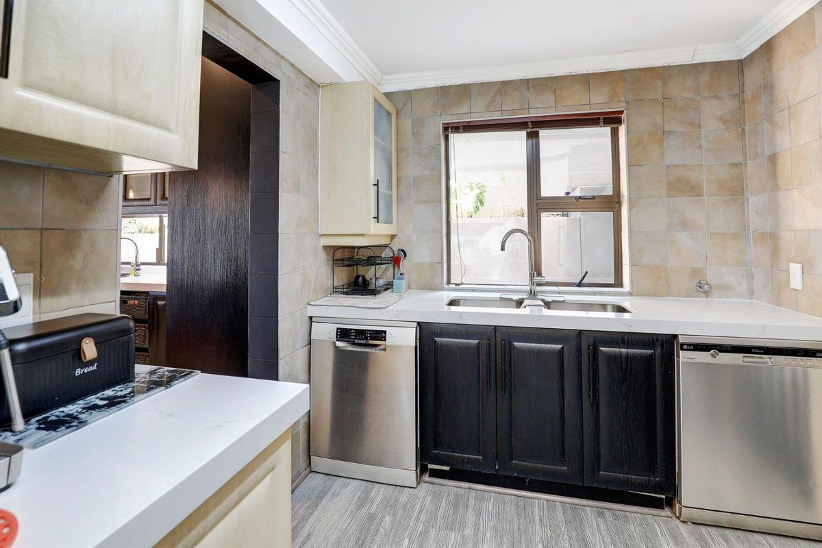 A functional kitchen featuring dark cabinetry, stone-look countertops, a double stainless steel sink, and integrated dishwasher space with neutral wall tiling.