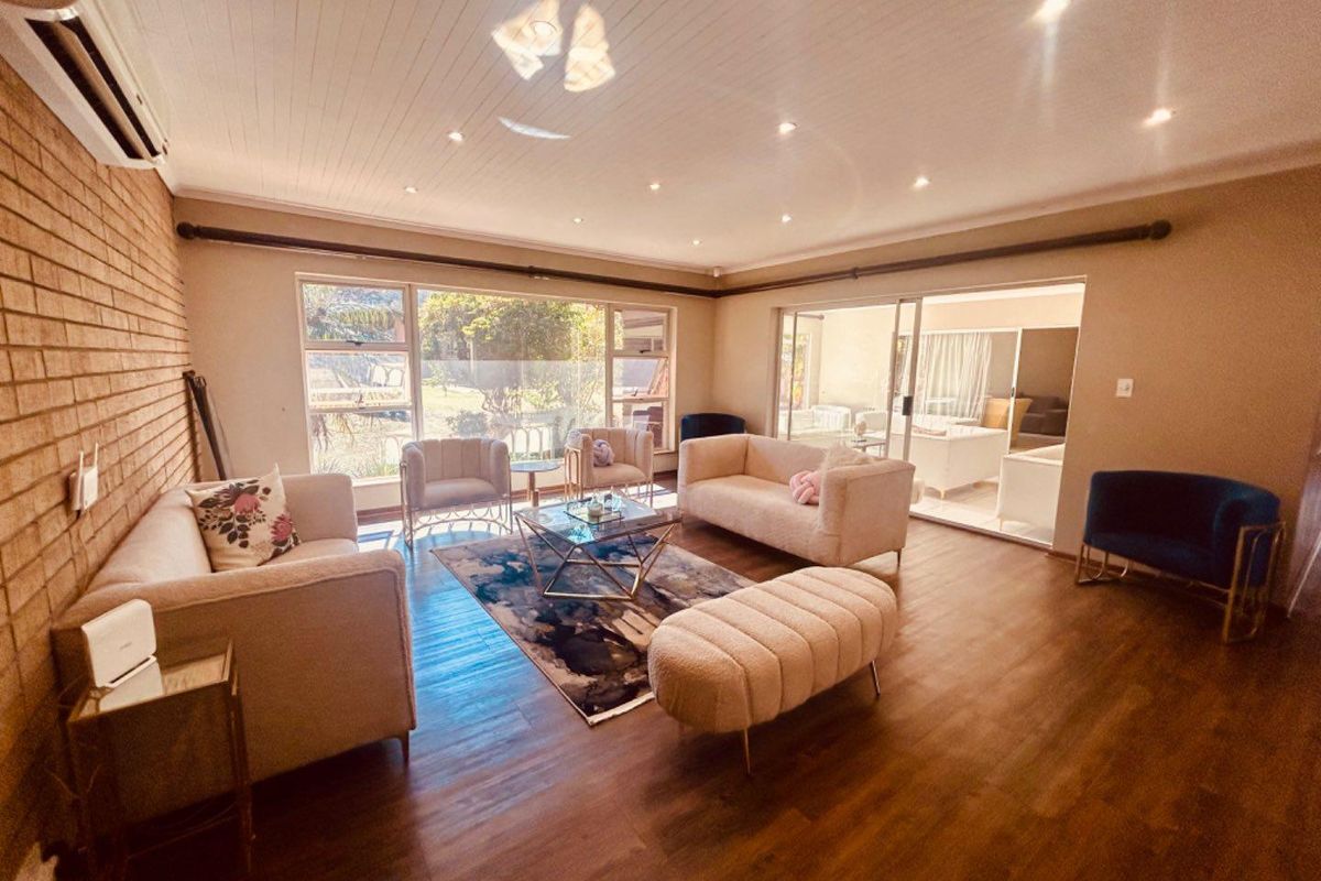 Spacious living room featuring wood-look flooring, a feature brick wall, recessed downlighting, and large windows providing natural light and access to the patio.