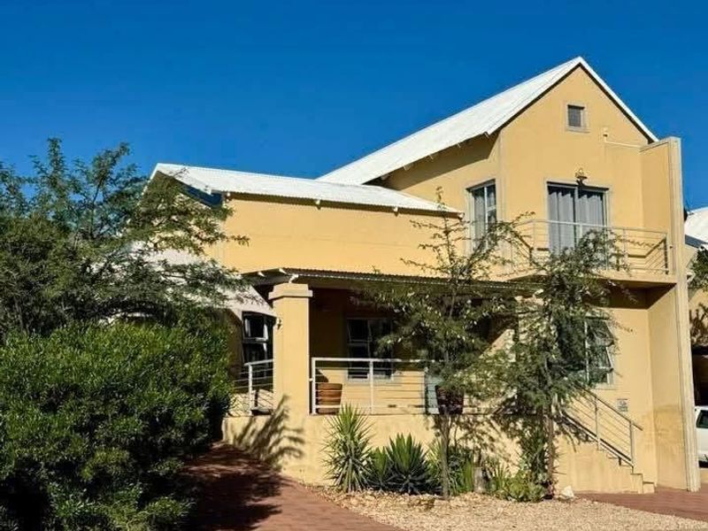 A two-story residential house featuring a yellow facade, corrugated metal roof, and an elevated balcony with a paved driveway and landscaped front garden.