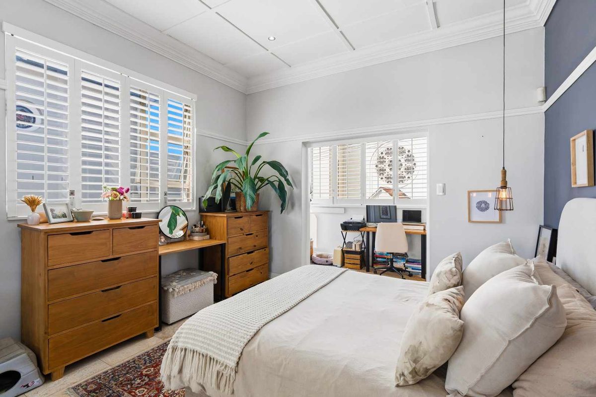 A well-lit bedroom featuring classic crown molding, a coffered ceiling, and plantation shutters, with an adjoining space suitable for a home office.