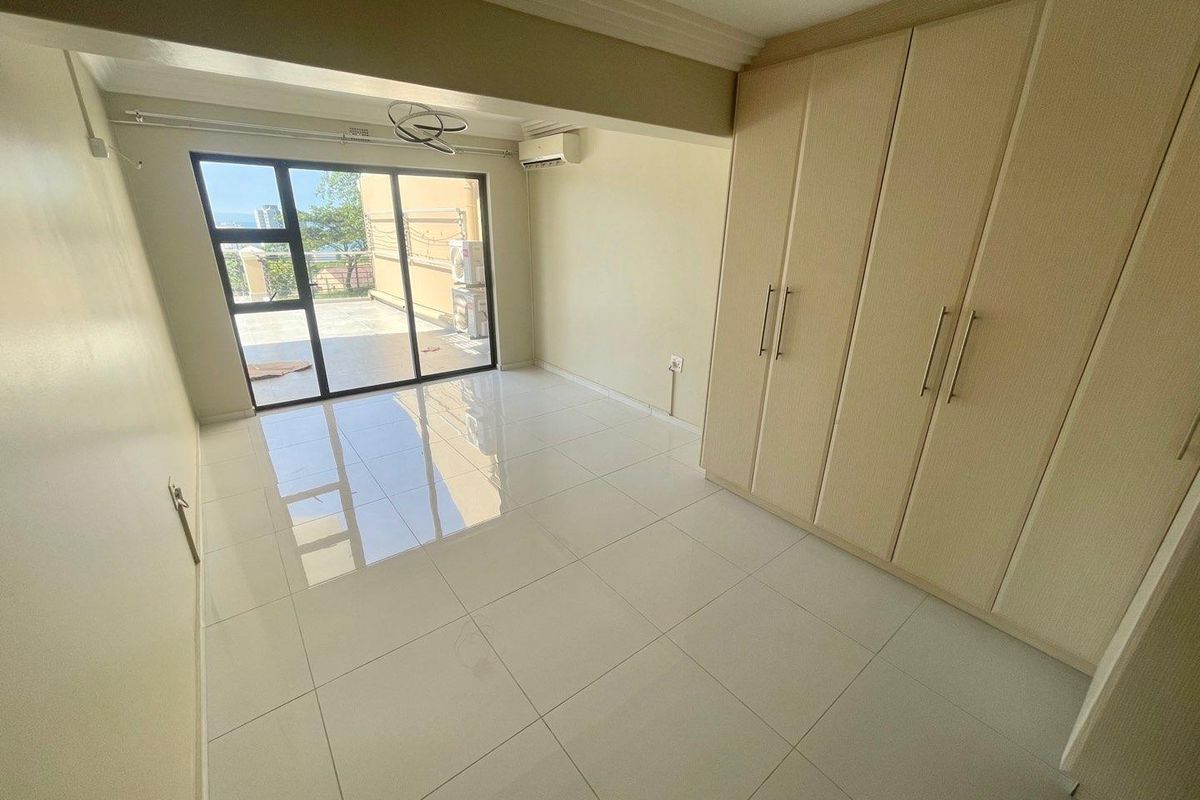 A bright, empty bedroom featuring light-colored floor tiles, built-in wooden wardrobes, and sliding glass doors leading to an outdoor balcony area.