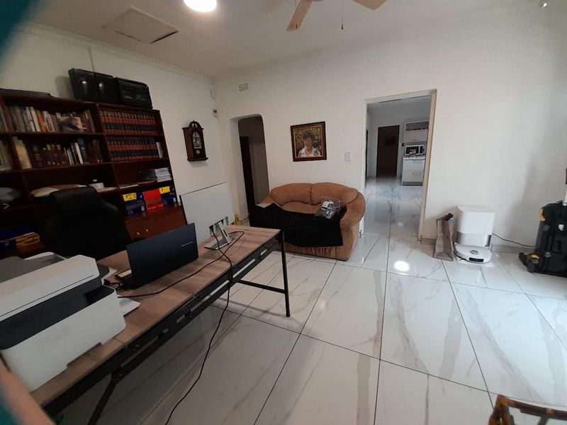 A spacious living area featuring large format white marble-look floor tiles, a ceiling fan, and an open archway leading to the kitchen area.