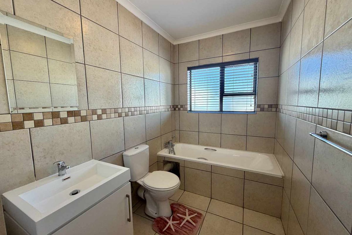 A clean, tiled bathroom featuring a built-in bathtub, a modern vanity with a basin, and a toilet, finished with neutral wall tiles.