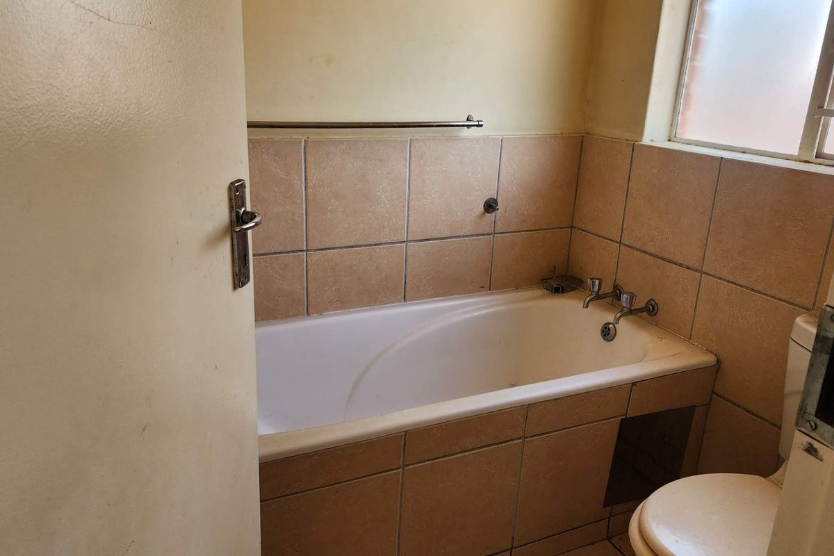 A dated bathroom featuring a built-in bathtub with tiled surround, a toilet, and a window with security bars.