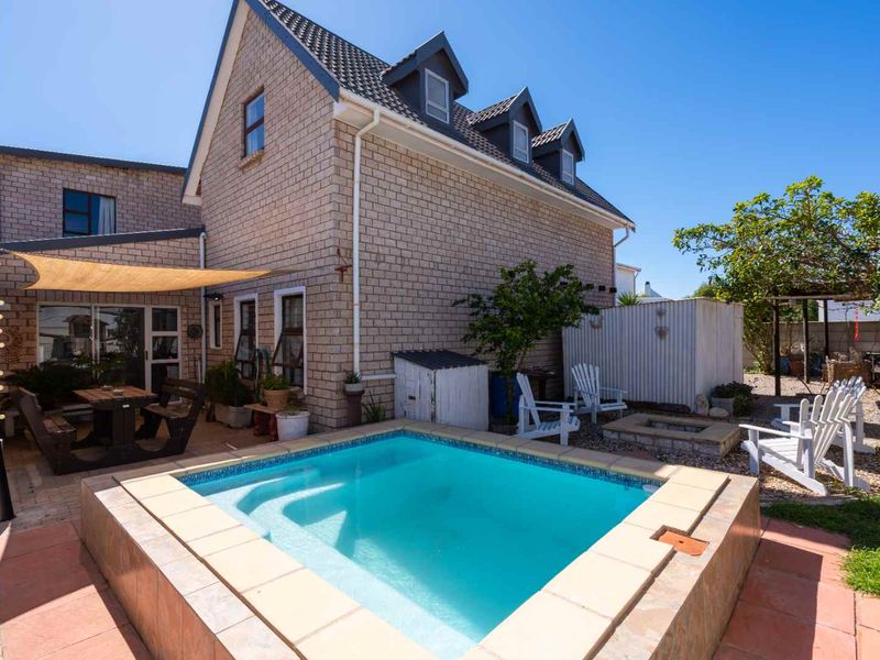 Rear exterior view featuring a swimming pool with tiled surround, paved patio area with shade sail, and brick house facade with dormer windows.