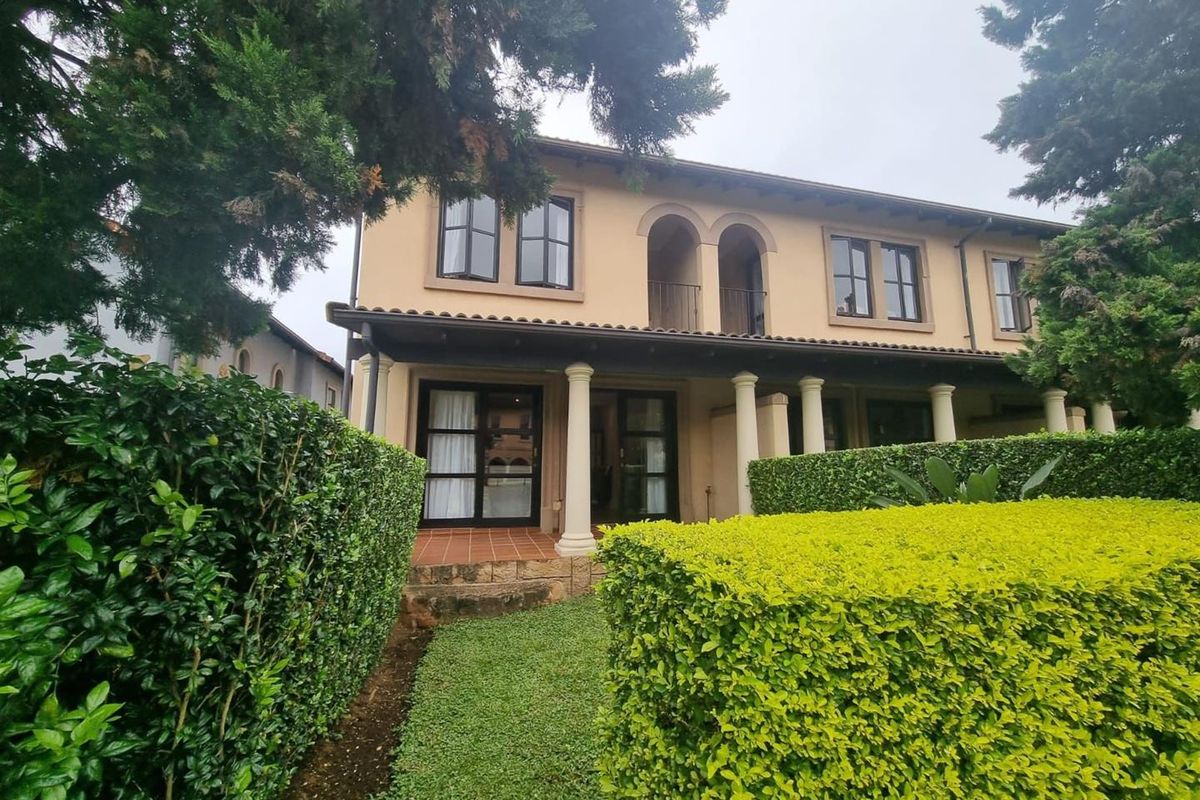 Rear view of a two-story townhouse featuring a covered patio with classical columns, a balcony with arched details, and well-maintained garden hedges.