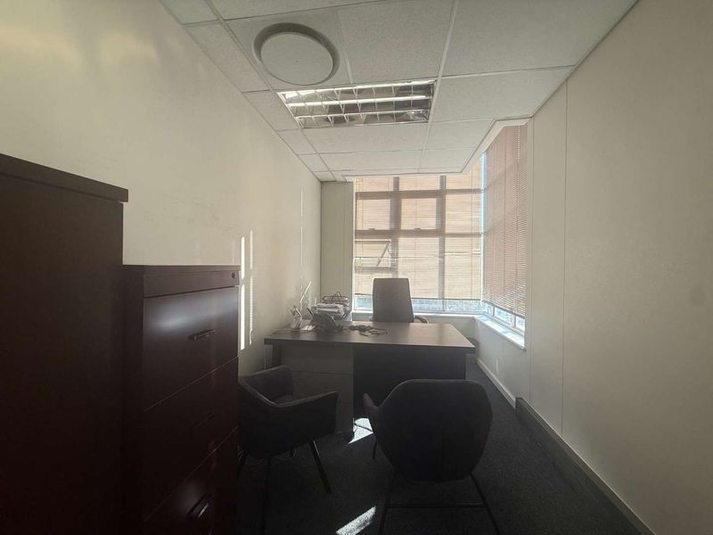 A functional office space featuring a suspended ceiling with recessed lighting, carpeted flooring, and large corner windows with venetian blinds for natural light control.