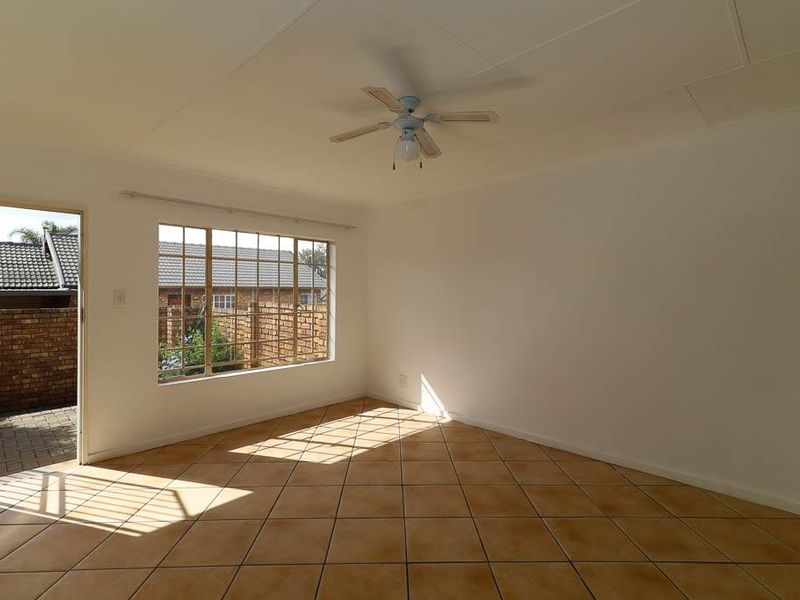 A bright, empty living room featuring neutral tiled flooring, a ceiling fan, and a large window providing natural light and access to the exterior.