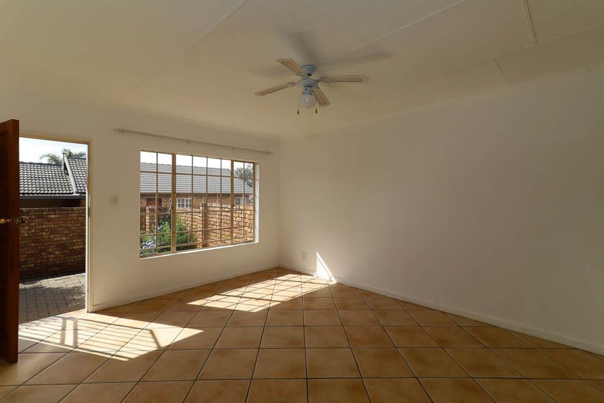 A bright, empty living room featuring neutral tiled flooring, a ceiling fan, and a large window providing natural light and access to the exterior.